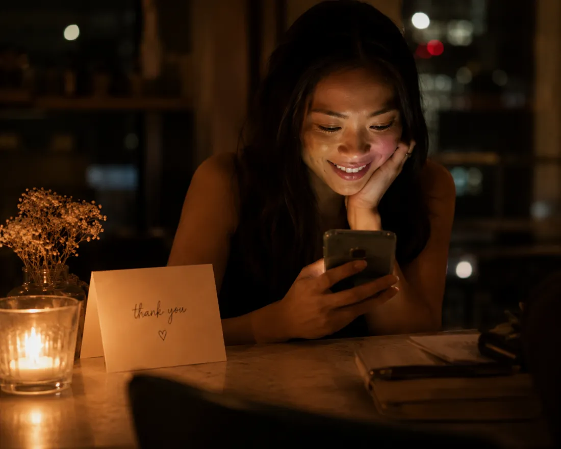 A woman smiling at her phone beside a handwritten thank-you card